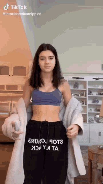 a woman in a blue bra and black sweatpants is standing in a living room with a white coat .