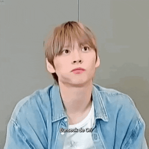 a young man wearing a denim jacket and a white t-shirt is making a funny face while sitting down .