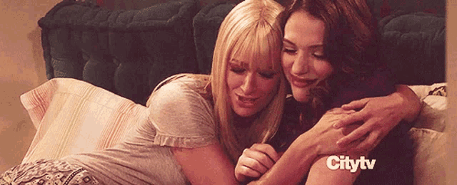 two women hugging on a bed with citytv written on the bottom right