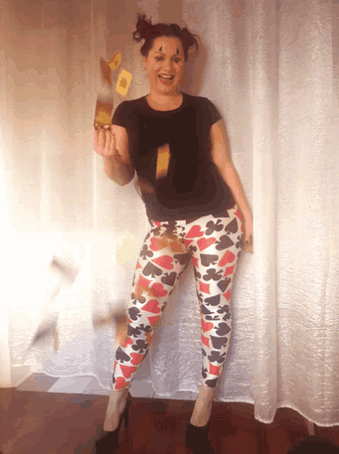 a woman wearing a black shirt and playing card pants holds a playing card in her hand