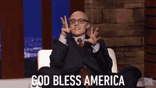 a man in a suit and tie is sitting in a chair and says god bless america