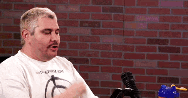 a man wearing a white shirt that says syndicate on it stands in front of a brick wall