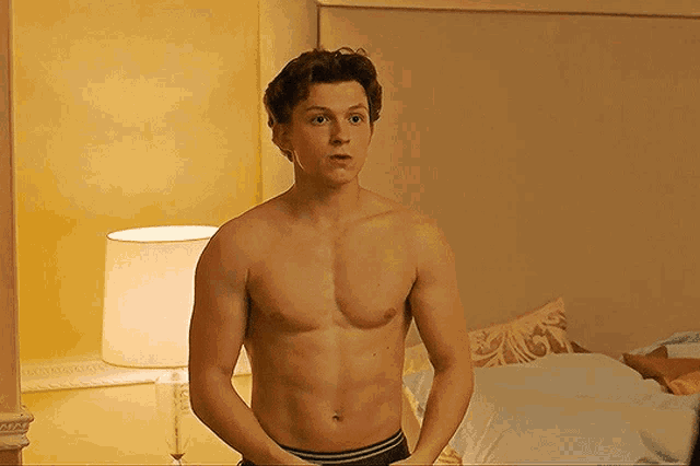 a shirtless man standing in front of a bed with a lamp in the background