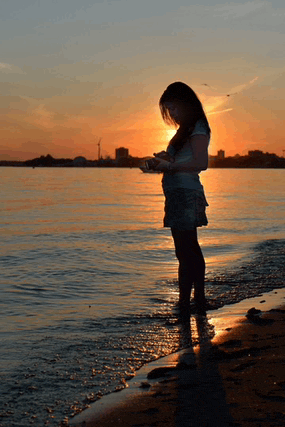 a woman standing on a beach looking at her phone at sunset
