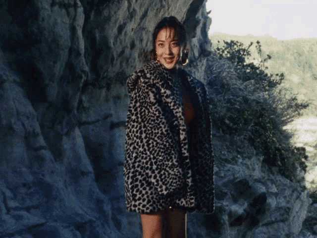 a woman wearing a leopard print coat stands in front of a rock wall