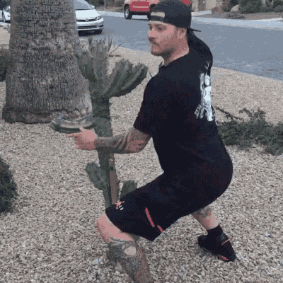 a man squatting next to a cactus wearing a black shirt with a skull on the back