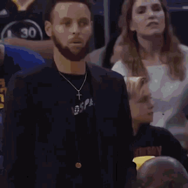 a man with a beard wearing a black jacket and a cross necklace is standing in a crowd of people .