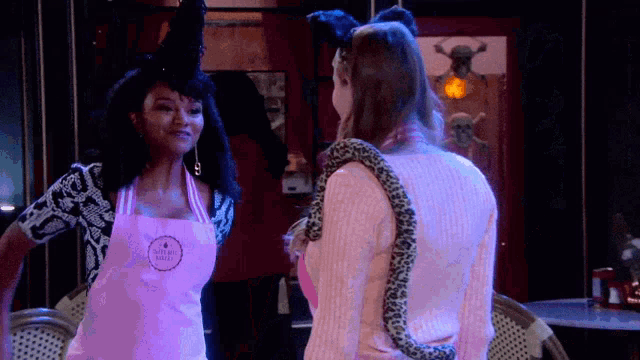 a woman wearing a pink apron that says hotel bakery stands next to another woman with a snake around her neck