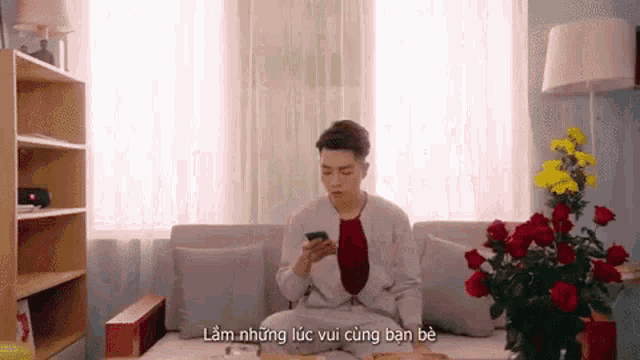 a man is sitting on a couch looking at his phone and flowers are in the background .