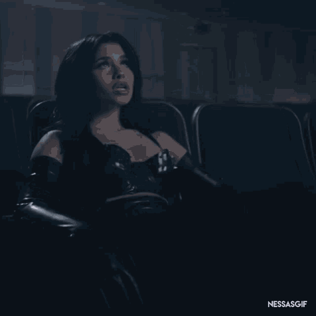 a woman in a black dress is sitting in a dark auditorium with the words nessasgif on the bottom right