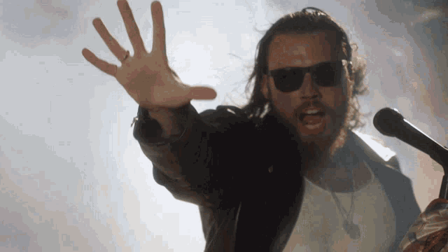a man wearing sunglasses and a leather jacket is singing into a microphone with his hand in the air