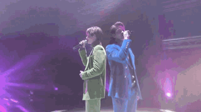 a man in a blue jacket sings into a microphone while another man in a green jacket looks on