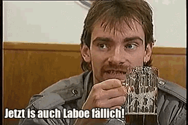 a man with a mustache is holding a glass of beer and says jetzt is auch laboe fallich .
