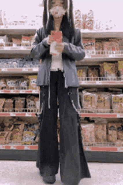 a woman wearing a mask is standing in front of a grocery store shelf holding a box .