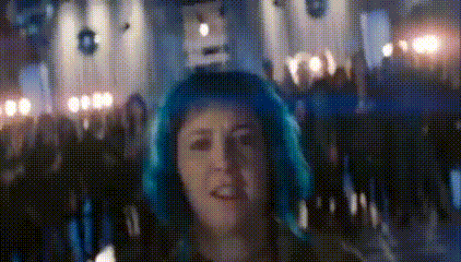 a woman with blue hair is standing in front of a crowd of people in a dark room .