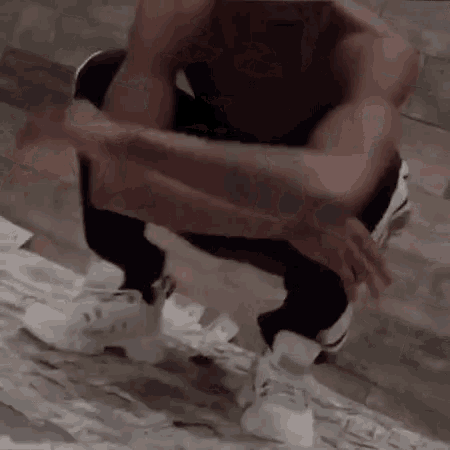a man without a shirt is squatting down on a wooden floor with his hands on his knees .