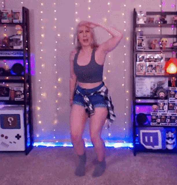 a woman in a crop top and shorts is dancing in front of a shelf with a twitch logo on it