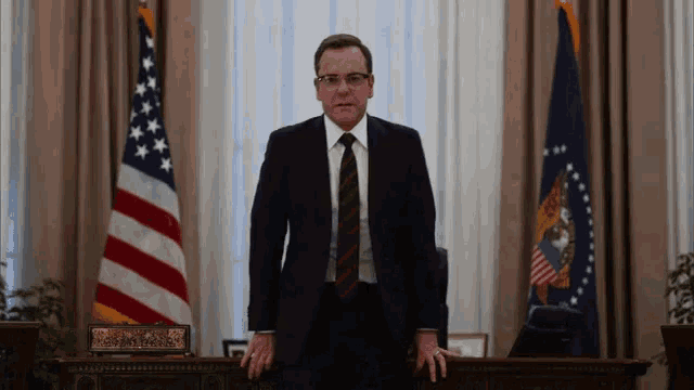 a man in a suit and tie is standing in front of a desk in a room with flags .