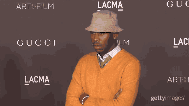 a man wearing an orange sweater and a tan hat is standing in front of a wall that says lacma