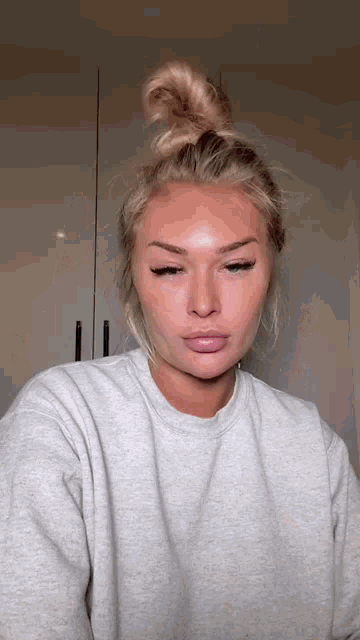 a woman with blonde hair in a bun is wearing a grey sweater and looking at the camera .