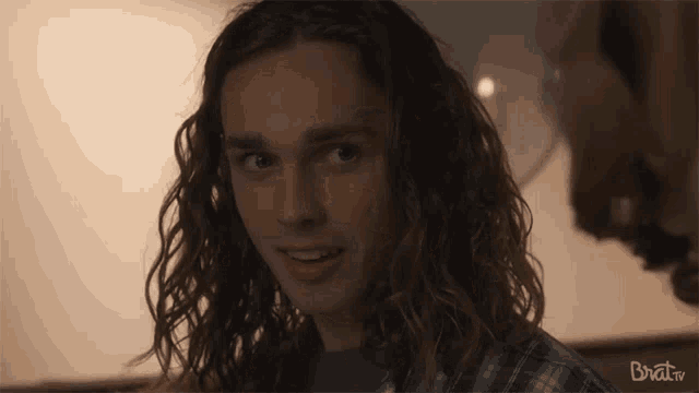 a young man with long curly hair looks at the camera with the words brat.tv visible in the corner