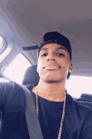 a young man wearing a hat and a necklace is taking a selfie in the back seat of a car .