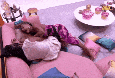 two women are laying on a pink couch in a living room with a bbc sign in the background