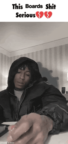 a man in a hooded jacket is sitting at a desk with a broken heart behind him that says this boards shit