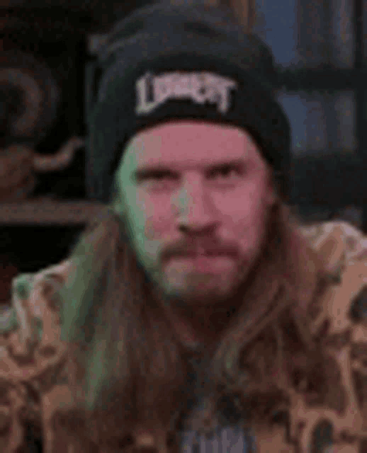 a man with long hair and a beard is wearing a black beanie and a camo jacket .