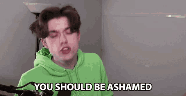 a young man in a green hoodie is standing in front of a microphone and says you should be ashamed .