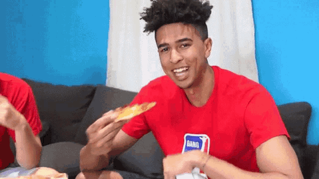 a young man in a red shirt is eating a slice of pizza while sitting on a couch .