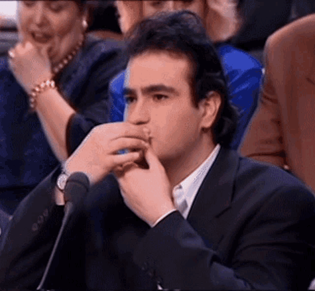 a man in a suit is sitting in front of a microphone and covering his mouth with his hand