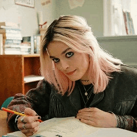 a woman with pink hair is sitting at a desk writing in a notebook