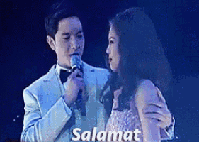 a man in a suit is singing into a microphone while a woman in a dress is hugging him .