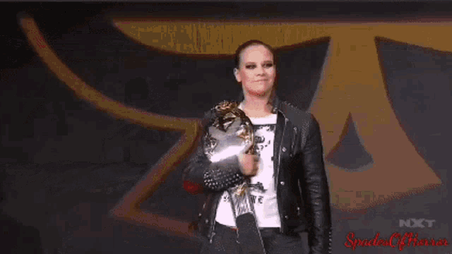 a woman in a black leather jacket is holding a trophy in front of a logo for nxt