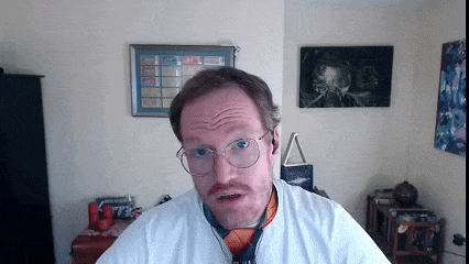 a man wearing glasses and a tie looks at the camera in front of a wall with a calendar on it