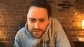 a man with a beard and a bun is sitting in front of a brick wall .