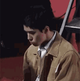 a man wearing a tan jacket and white shirt is sitting on a red carpet