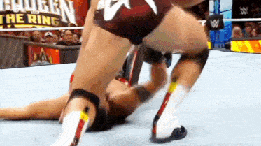 a wrestler is laying on the ground while another wrestler is standing on top of him .