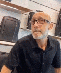 a bald man with glasses and a beard is sitting in front of a display of suitcases .