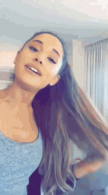 ariana grande is wearing a blue tank top and a ponytail while standing in a room .