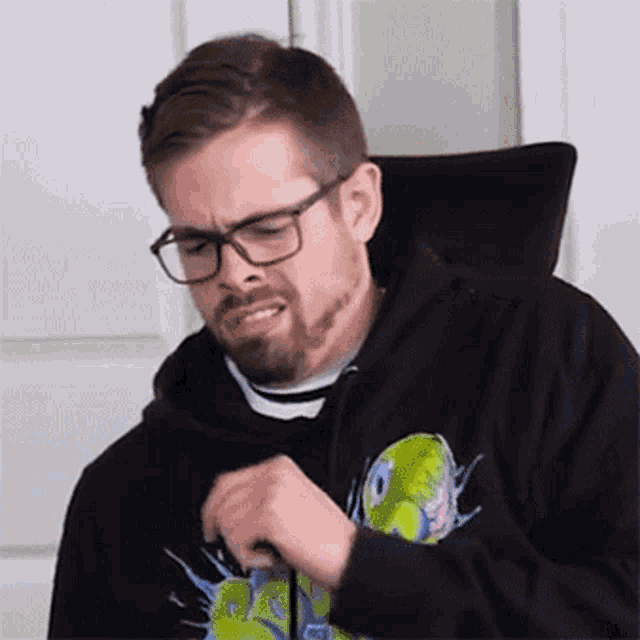 a man with a beard and glasses is wearing a black hoodie and making a funny face .
