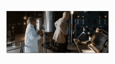 a man in a lab coat talks to another man in a dark room