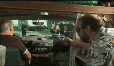 two men are sitting in a car talking to each other and looking out the window
