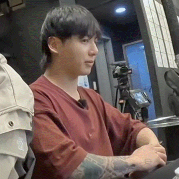 a man with a tattoo on his arm is sitting at a table with a camera in the background .