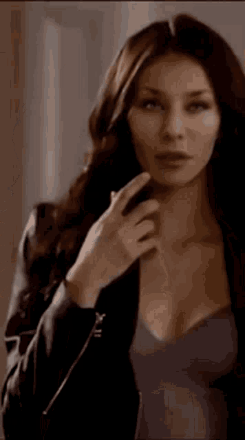 a woman with long brown hair is wearing a black jacket and a gray top .