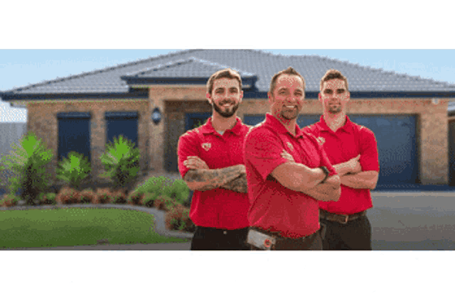 three men in red shirts standing in front of a house with their arms crossed