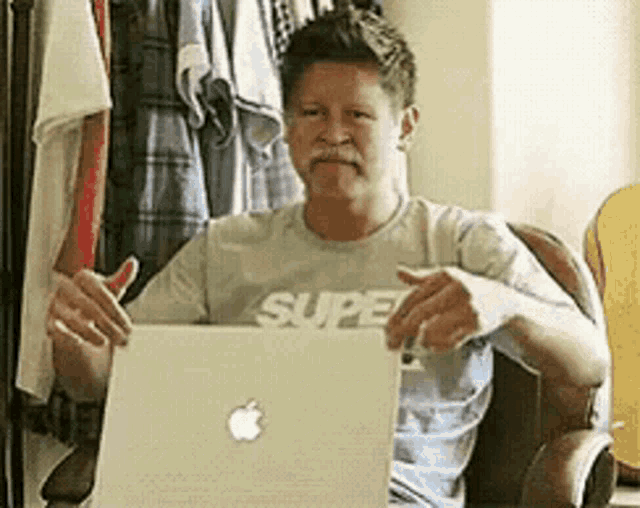 a man wearing a shirt that says super is sitting at a desk with an apple laptop