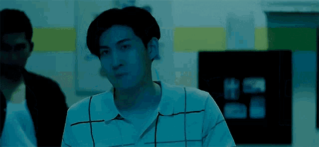a man in a blue shirt is sitting in a dark room with a man in a black jacket standing behind him .