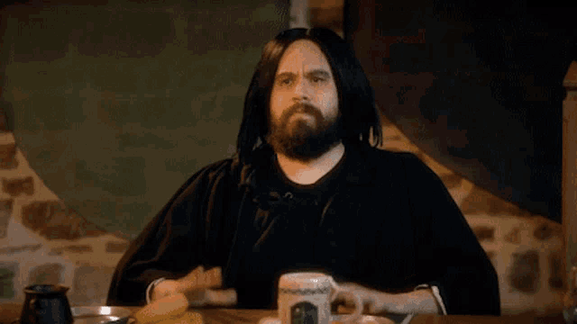 a man with long hair and a beard is sitting at a table with a cup of coffee